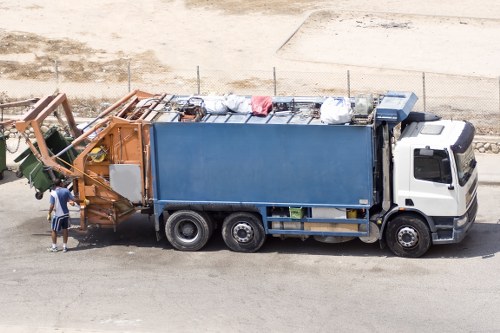 A large commercial rubbish collection truck with a white cab and a blue metal waste compartment is parked on a paved road in an outdoor area. The truck is loaded with various waste materials including white plastic bags, cardboard boxes, and metal items stacked along the top of the container. In front of the truck, a person wearing casual clothing and a cap is standing next to the waste disposal area, possibly overseeing or assisting with the rubbish loading process. The background shows a sandy or gravelly surface, a simple wire fence, and an open area that suggests a site for waste collection or disposal. The scene is well-lit, indicating daytime, and the environment appears clean and orderly, consistent with rubbish removal operations in the Wimbledon area as managed by Business Waste Removal Wimbledon. The truck's design and the waste materials reflect typical commercial waste collection services focused on rubbish removal in London suburbs.