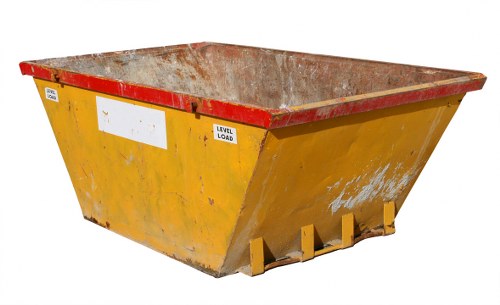 A large, rectangular industrial skip with a yellow painted exterior and a red top edge, positioned outdoors on a paved surface. The interior of the skip reveals a rusty, textured metal surface indicating prior use for waste collection. The skip is supported by metal lifting points at the front, with their yellow paint slightly worn. The background shows an outdoor environment with no specific surroundings visible. The skip appears to be ready for waste removal, typical of commercial or construction rubbish disposal services provided by companies such as Business Waste Removal Wimbledon, serving local areas including Wimbledon and surrounding postcode districts in south-west London. The overall scene highlights the utilitarian design of waste containers used for large-scale rubbish collection and removal, in a setting appropriate for professional waste management services.