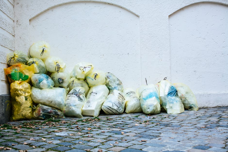 A collection of tightly packed transparent plastic rubbish bags filled with waste, including household debris, stacked against a white textured wall on a cobblestone pavement. The bags vary in size an